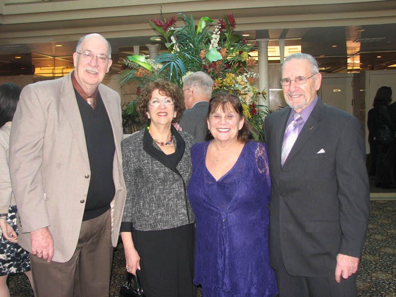Four people pose together near a floral arrangement. Two men in suits, two women in dresses, all smiling.