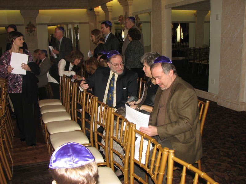 People, some wearing yarmulkes, in a room, looking at papers. Chairs are in rows.