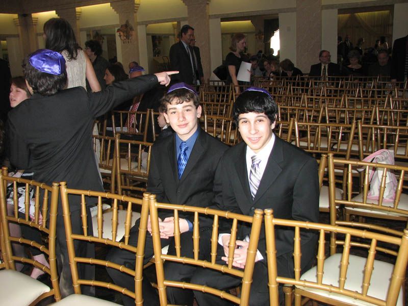 Two boys in suits and yarmulkes sit in golden chairs at an event. A person points.