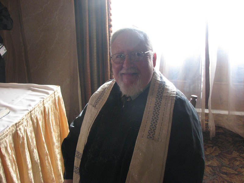 Man with a white beard and glasses, wearing a black shirt and a shawl, sitting near a window with sunlight.