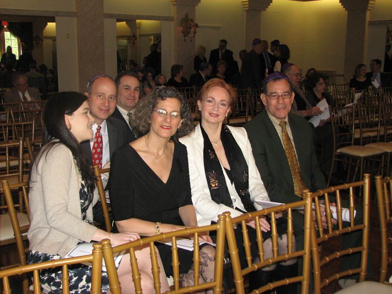 People seated at an event, likely a ceremony. They sit on gold chairs, with others in the background.