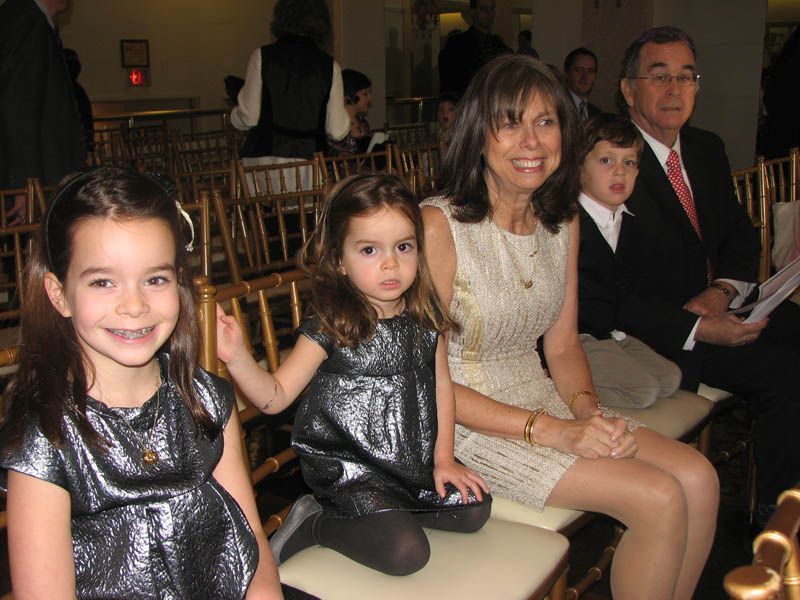 Family seated at an event. Girls in silver dresses, woman in cream dress, man in suit. Indoors, gold chairs.