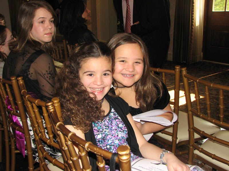 Two girls smile at the camera while seated in gold chairs at an event.