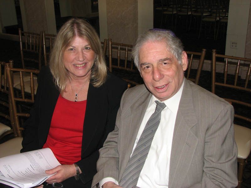 Woman in red top and man in suit seated, both smiling, inside a room.