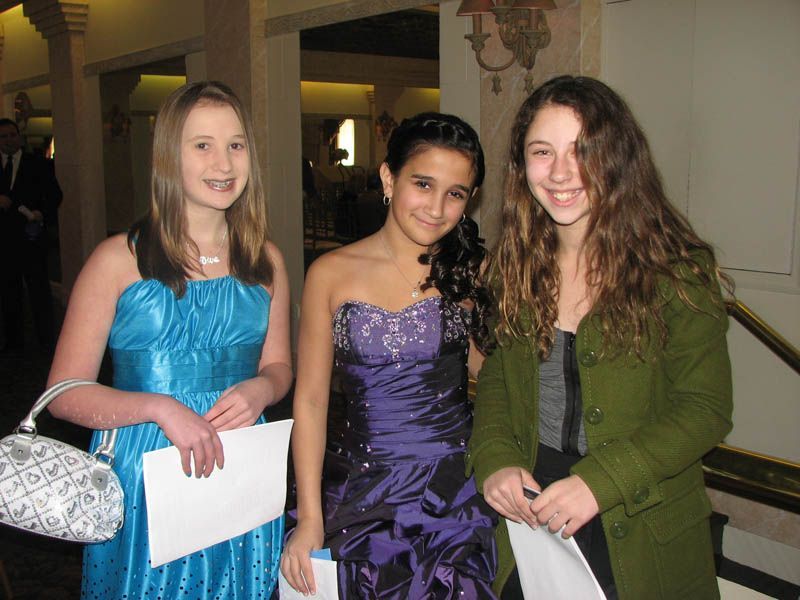 Three girls pose indoors. One in blue dress, one in purple dress, and the third in a green coat. All hold papers.