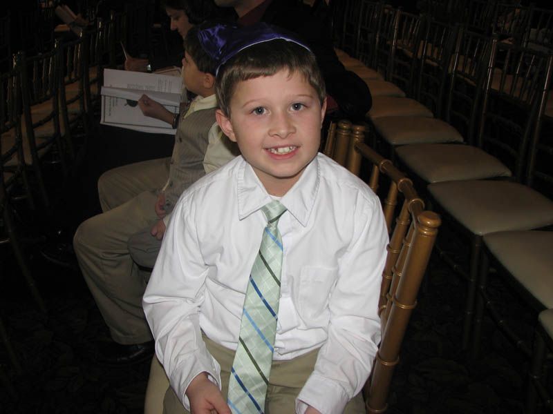 Boy wearing a yarmulke and tie, sitting in a chair at an event.