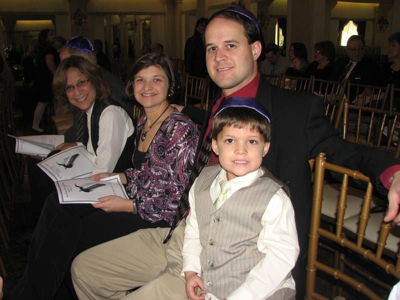 Family seated, wearing yarmulkes, at an event. The boy in a vest smiles, others hold programs.