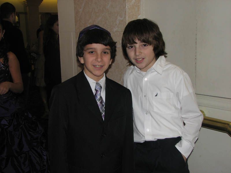 Two boys in formal attire smiling near a wall. One in a suit and kippah, the other in a button-down shirt.