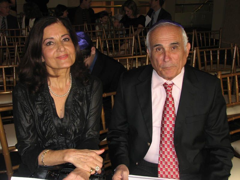 Couple seated at an event, woman with dark hair, man with yarmulke, both dressed formally.