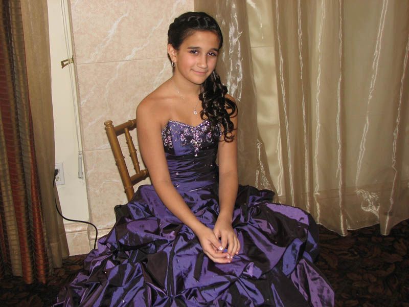 Woman in a purple formal gown sits on a chair. Dark hair is pulled to the side. Behind her are gold drapes.