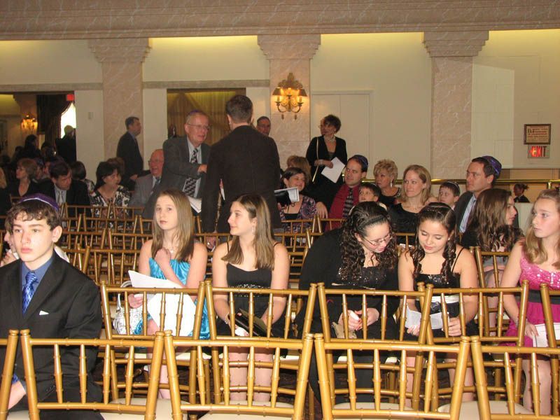 People seated at an event, many wearing formal attire. Golden chairs fill the room, pillars in the background.