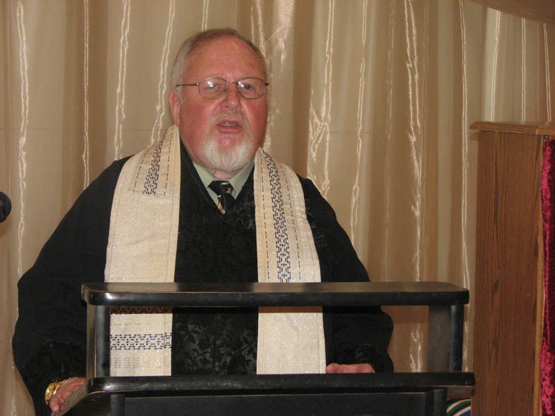 Man in glasses and clerical garb speaks from a podium, white scarf with black design.