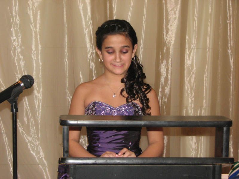 Girl in purple formal dress, at podium, with microphone, curtains in background.