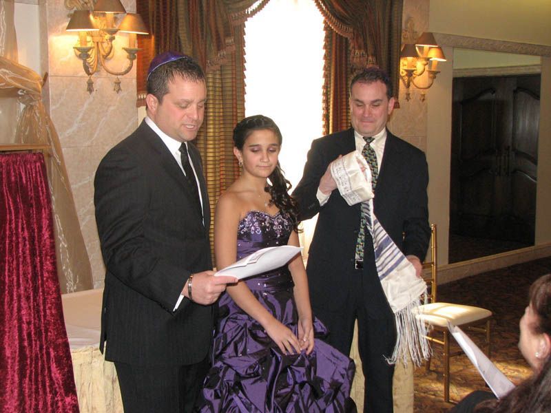 A girl in purple gown at a bar mitzvah ceremony; two men in suits, one reading, one holding a tallit.