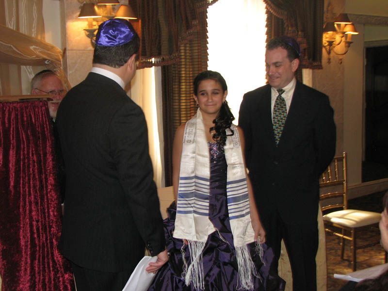 A girl in a purple dress and tallit stands with two men during a Jewish ceremony.