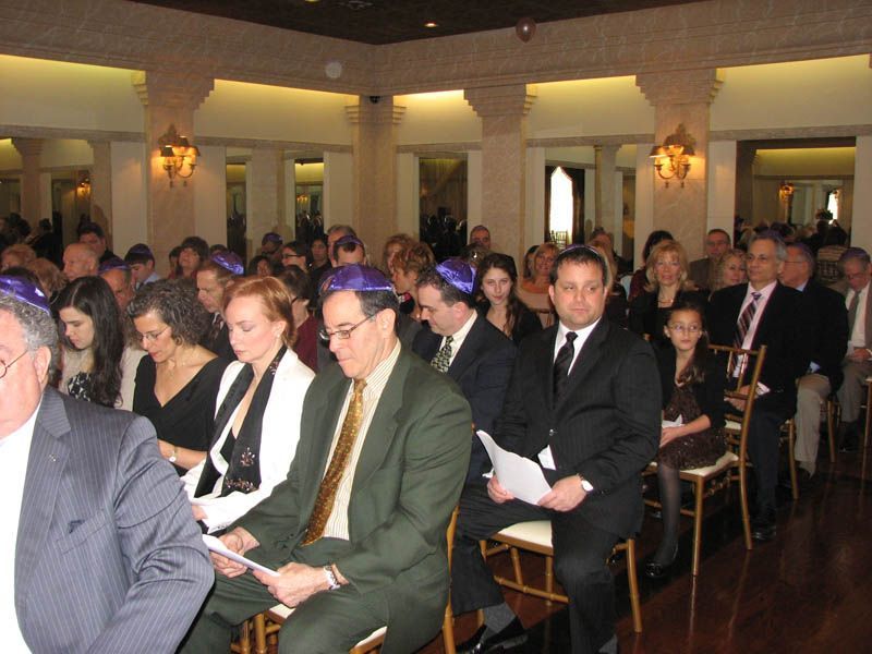 People in a formal room, many wearing yarmulkes, seated and listening.