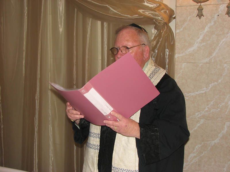 Man in glasses wearing a tallit and reading from a pink folder. Beige curtain background.