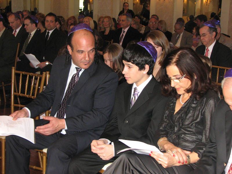Family at a Jewish ceremony; man, boy, and woman seated, wearing kippahs, looking at papers.