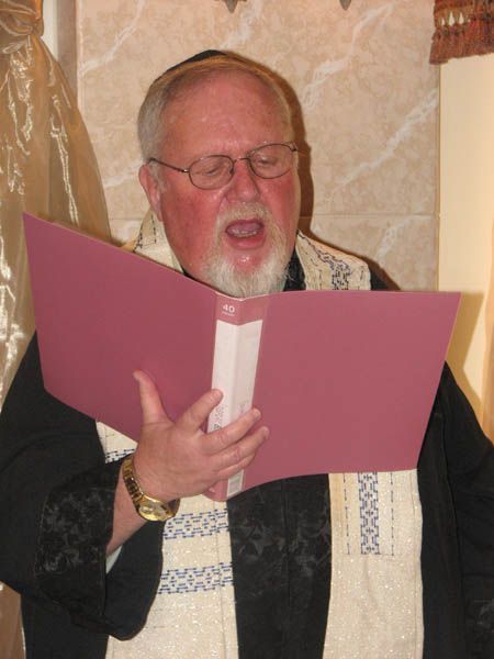 Man in yarmulke and tallit singing from a pink book, indoors.