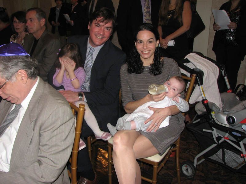 Family at an event; mother feeding baby, father and daughter sitting. Formal setting, smiling.