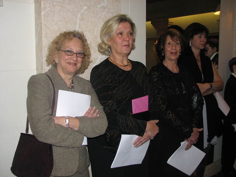 Three women stand together, holding papers, in an interior setting. One has arms crossed. All appear to be observing.