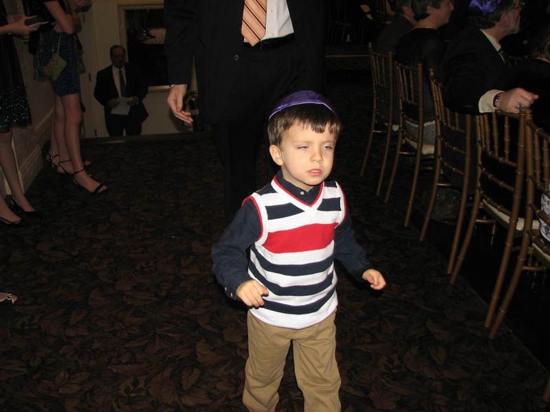 Boy in yarmulke dances, wearing sweater vest and khaki pants at event.