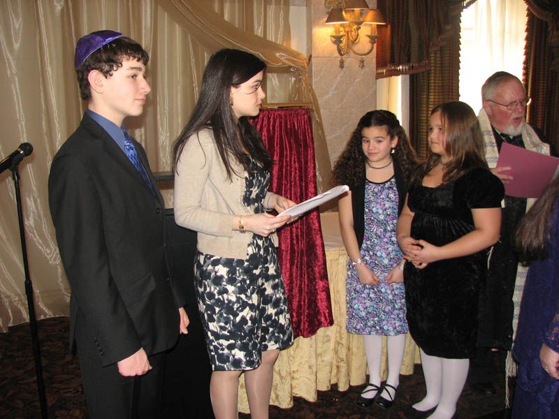Bar Mitzvah ceremony: person reads, others stand nearby, rabbi officiates. Indoors, festive setting.
