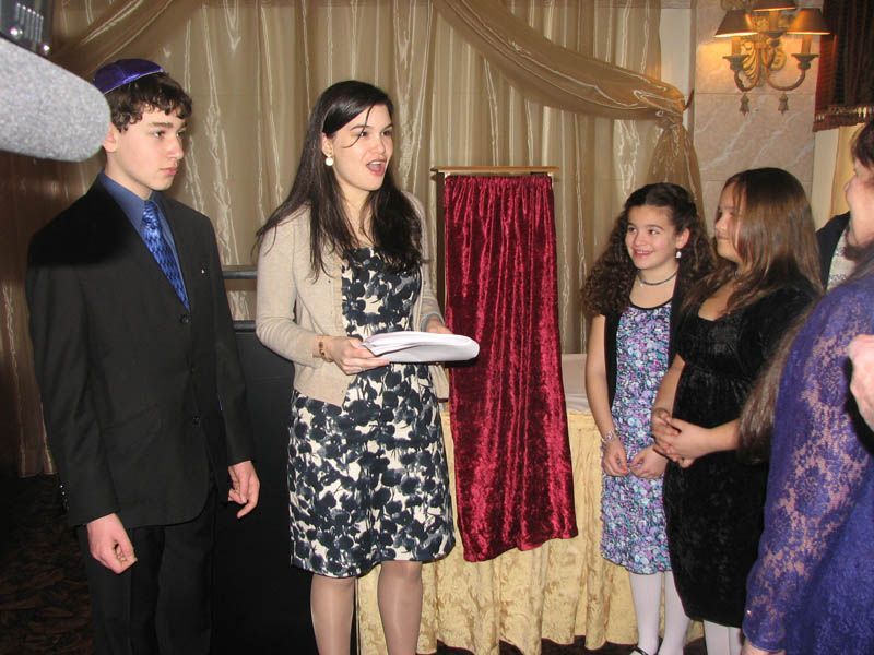 A woman speaks to a group at an event; a boy in a suit, and girls in dresses listen. Gold decor in background.