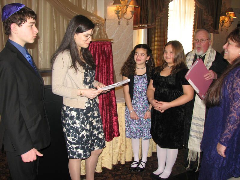 A Jewish ceremony: a woman reads, a boy in a yarmulke and two girls stand, a rabbi and woman watch.