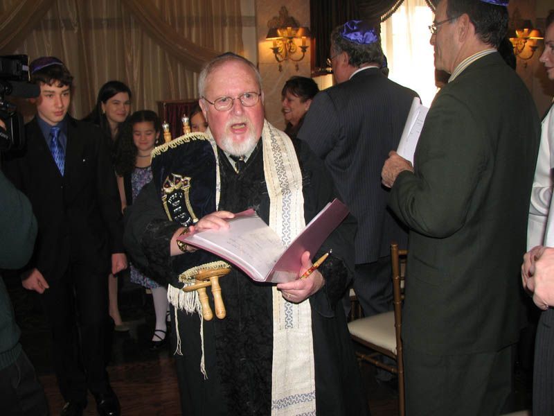 Man in religious garb reads from a book, surrounded by people in an indoor setting.