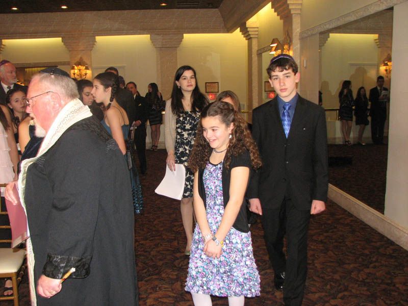 People walking through a formal hall. Several are dressed in suits and dresses. An older man wears a religious robe.