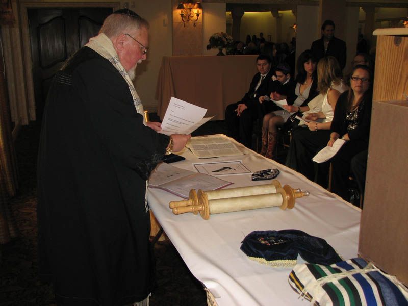 A rabbi reads from a paper during a Jewish ceremony, with a Torah scroll on a table and guests seated in the background.