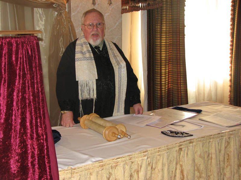Man wearing a tallit stands behind a table with a Torah scroll.