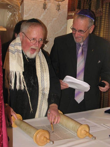 Two men read a Torah scroll at a table. One wears a tallit, the other a yarmulke.