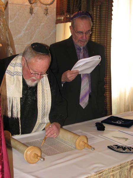 Two men reading a Torah scroll. One wears a tallit and kippah, the other a kippah and suit. Indoors, at a table.