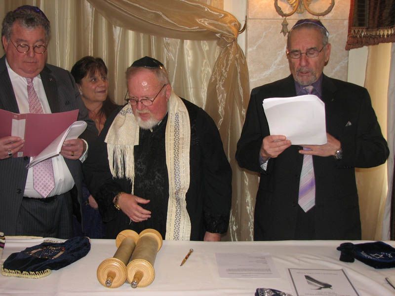 Three men reading papers at a table with Torah scrolls. A woman looks on.
