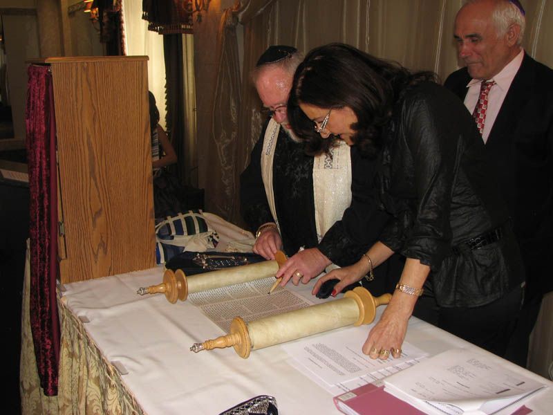 People signing a document on a table with a Torah scroll. A man and a woman are writing, another man watches.