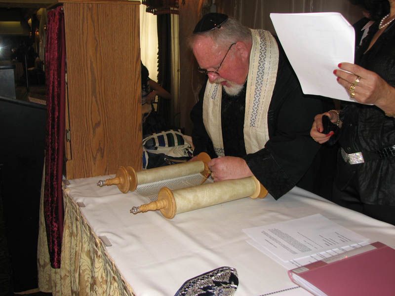 Man in black robes, yarmulke, and tallit, reads Torah scroll on table, possibly at a ceremony.