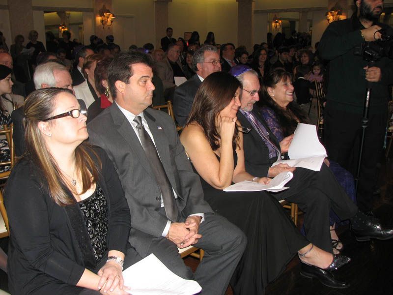 People seated at an event, some holding papers. Dark attire, indoors with ornate decor.