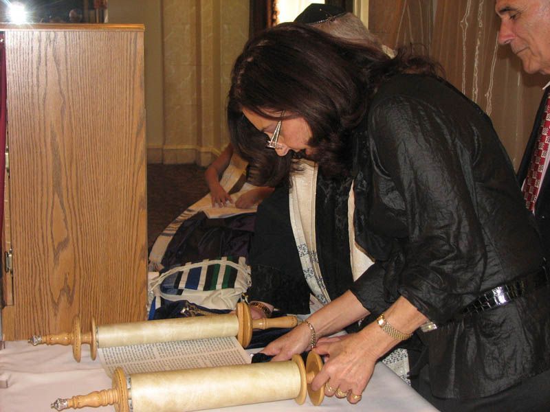 Woman rolling a Torah scroll in a synagogue, with a wooden cabinet and another person present.