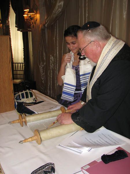 A person in a tallit and kippah, with a young person, opening a Torah scroll on a table.