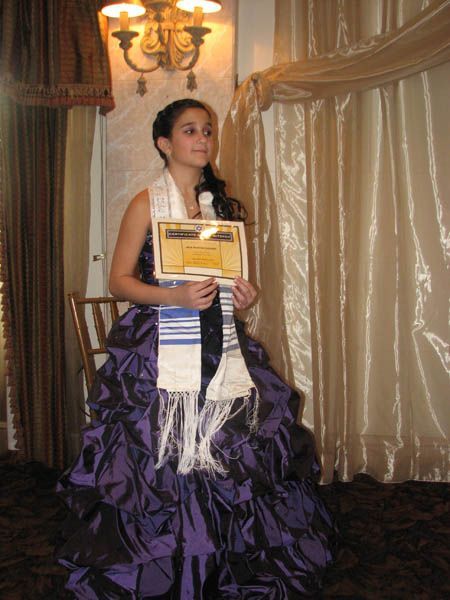 Young person in purple gown holding a framed document, wearing a prayer shawl, at an event.