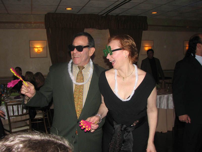 Two people at a party: man with sunglasses and lei, woman with shamrock glasses, both holding noisemakers.