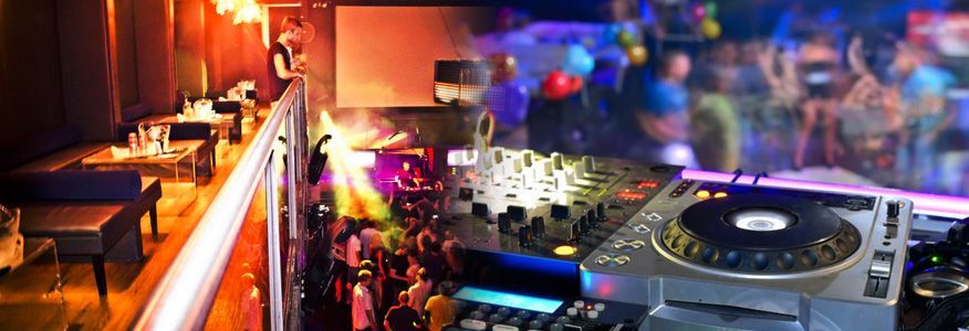 DJ mixing music in a club with people dancing and a seating area with tables and a balcony.