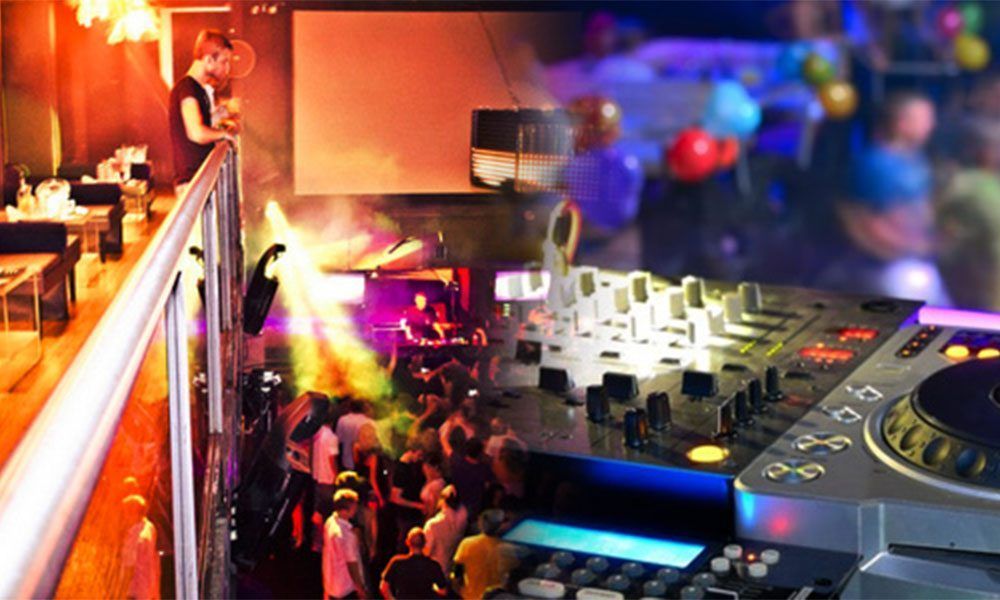 Party scene: DJ setup, dancing crowd, upper level with bar and person, colorful lights.