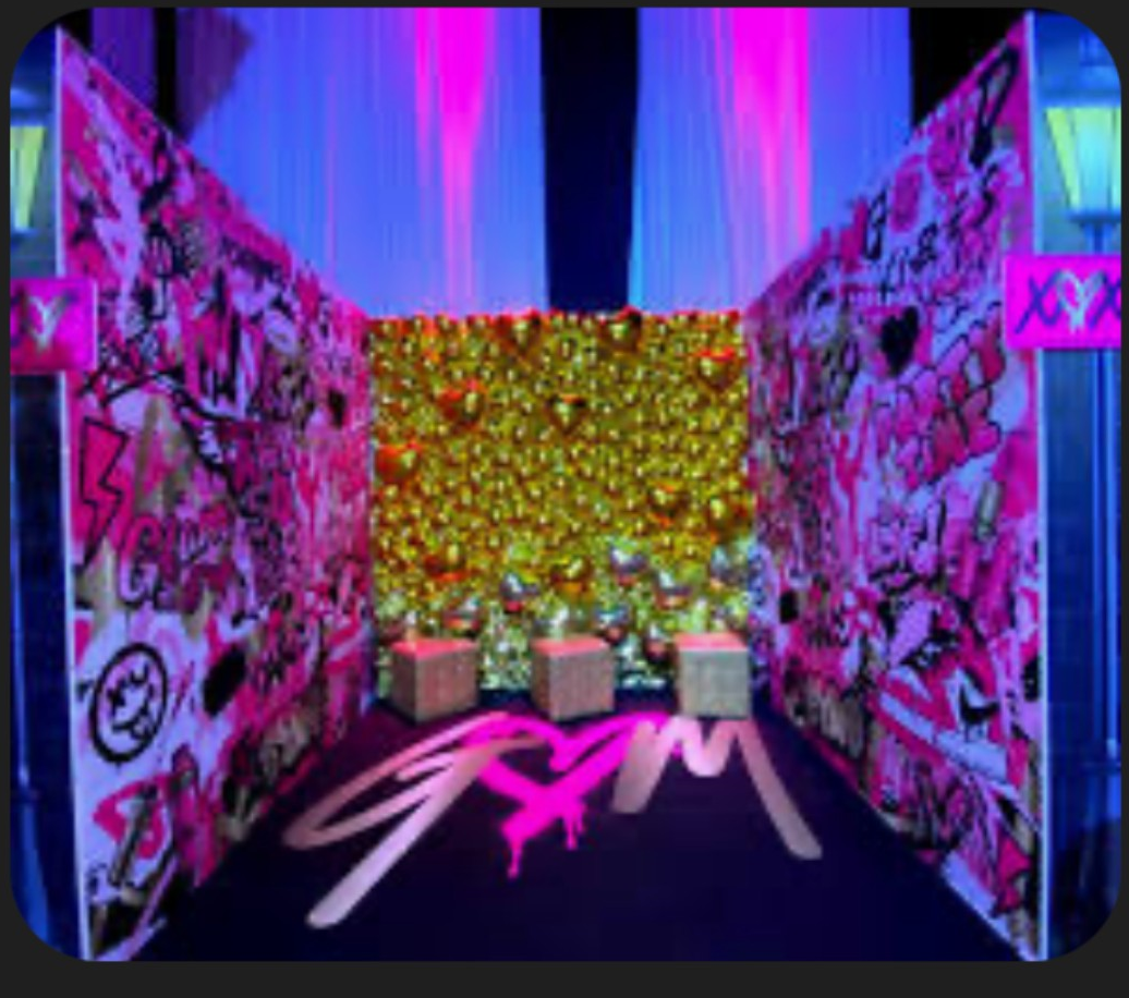 Room with graffiti-covered walls, gold balloon backdrop, three cubes, and neon pink and blue lighting.