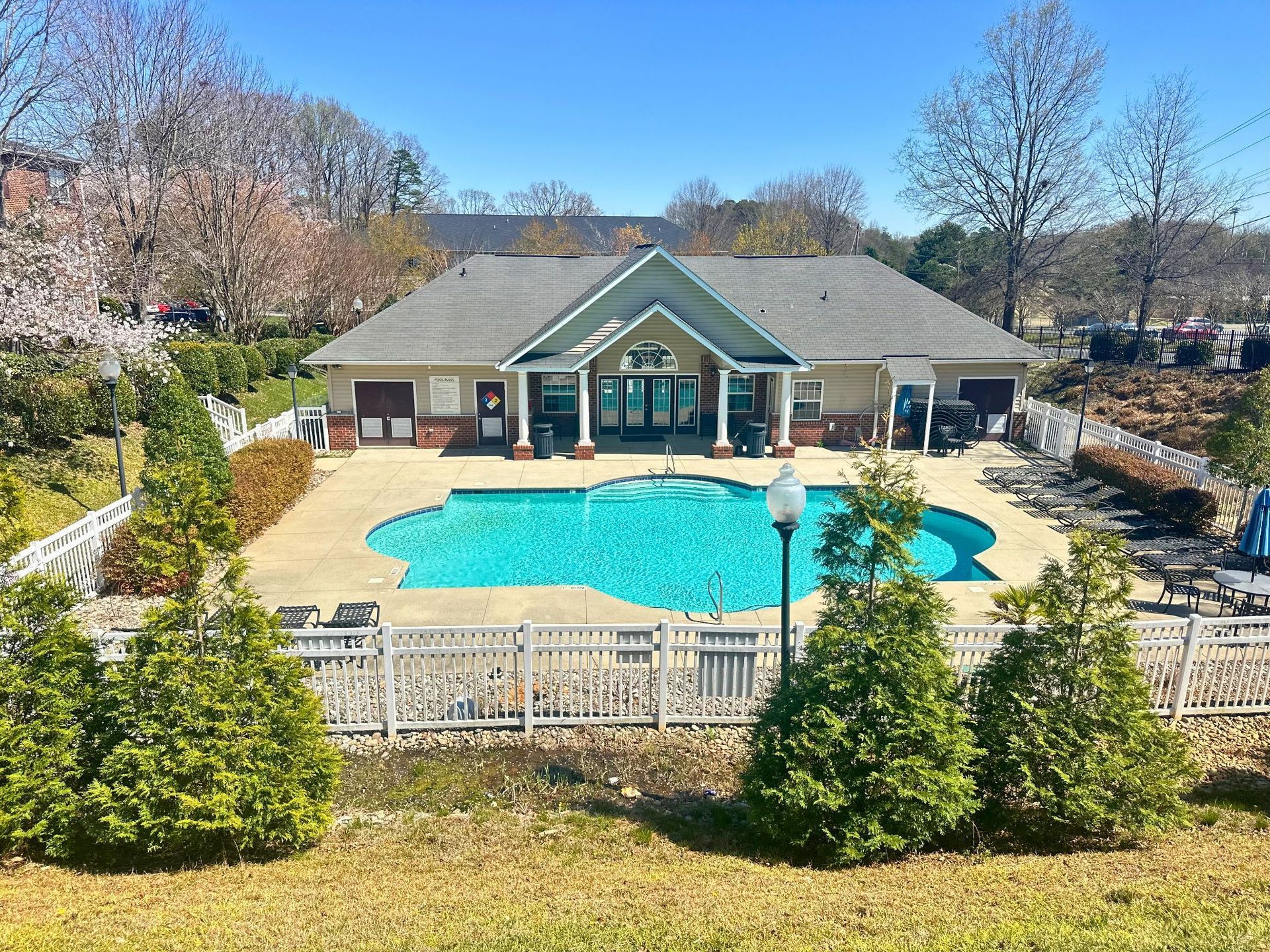 A pool with turquoise water in front of a clubhouse with a gray roof, surrounded by landscaping.