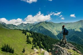Hiker with backpack standing on a rocky mountain ridge overlooking green valleys and distant peaks under a blue sky