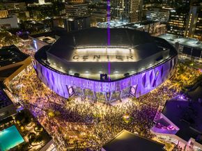 Nighttime aerial of a packed, purple-lit arena surrounded by city buildings and crowds