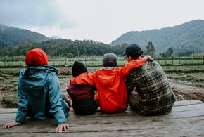 Four people sit on a wooden deck facing misty green hills, one in a red hooded jacket.